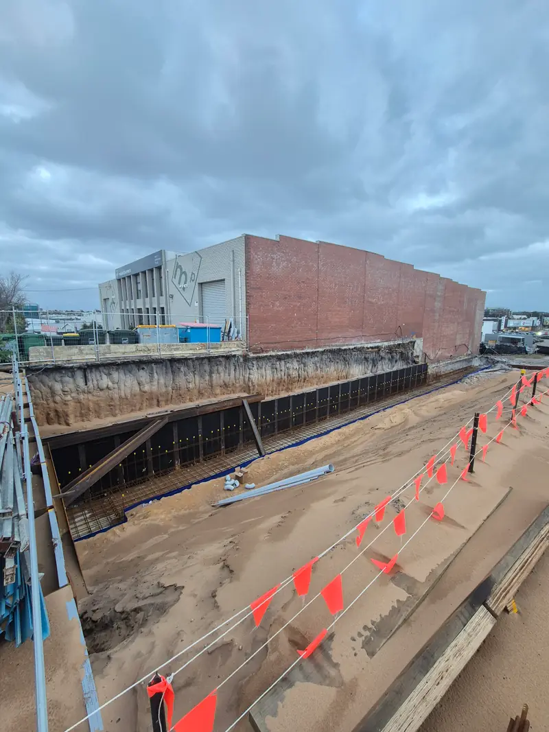 Large commercial excavation with steel bracing and safety fencing beside a brick warehouse